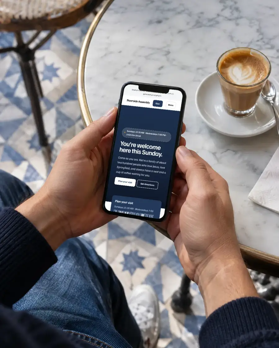 Illustrative photograph of hands holding a phone showing an example church website at a café table.